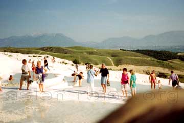 Touristen beim baden in den seichten natürlichen Becken auf den Kalkterrassen von Pamukkale in Kleinasien, Türkei. Im Hintergrund ein See und ein hohes mit Schnee bedecktes Gebirge.